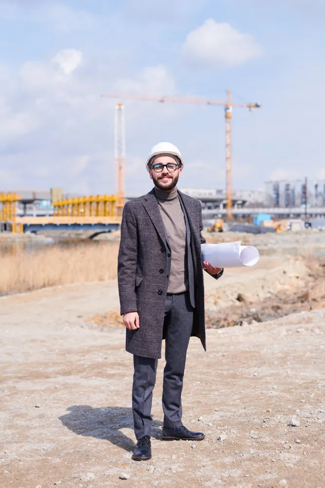 Civil engineer wearing a safety helmet holding construction blueprints at an active construction site with cranes in the background